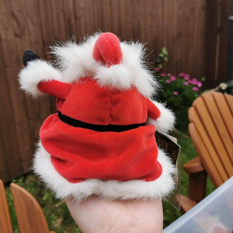 K&ouml;nnte beinhalten: Ein rotes Pl&uuml;sch-Weihnachtsmann-Spielzeug mit wei&szlig;em Pelzbesatz und einem schwarzen G&uuml;rtel. Das Spielzeug h&auml;lt ein schwarzes Objekt in seiner linken Hand.