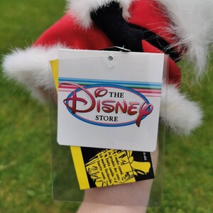 K&ouml;nnte beinhalten: Ein gelbes Etikett mit dem Text "THE DISNEY STORE" in einem roten Kreis. Das Etikett ist an einem Pl&uuml;schtier mit einer rot-wei&szlig;en Weihnachtsm&uuml;tze befestigt.