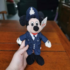 K&ouml;nnte beinhalten: Ein Pl&uuml;sch-Mickey-Mouse-Spielzeug, das eine blaue Polizeiuniform mit einem schwarzen Hut und einem wei&szlig;en Etikett mit der Aufschrift "The Disney Store" tr&auml;gt.