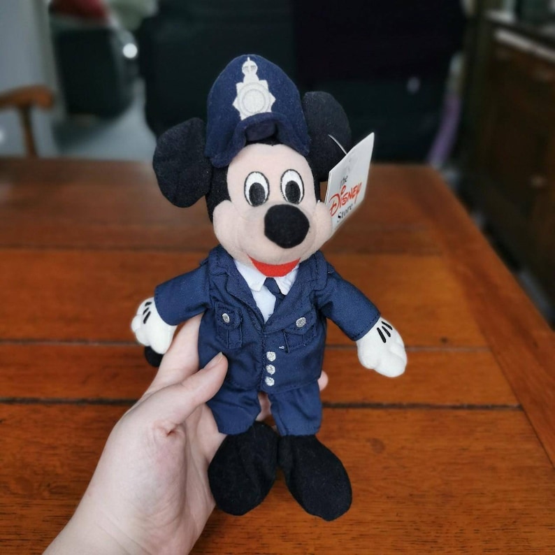K&ouml;nnte beinhalten: Ein Pl&uuml;sch-Mickey-Mouse-Spielzeug, das eine blaue Polizeiuniform mit einem schwarzen Hut und einem wei&szlig;en Etikett mit der Aufschrift "The Disney Store" tr&auml;gt.