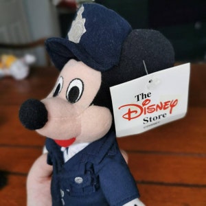 K&ouml;nnte beinhalten: Ein Pl&uuml;sch-Mickey-Mouse-Spielzeug, das eine blaue Polizeiuniform mit einem silbernen Abzeichen tr&auml;gt. Das Spielzeug hat ein schwarzes Etikett, das an seinem Ohr befestigt ist und "The Disney Store" mit einem Copyright-Symbol und "Disney" darunter tr&auml;gt.