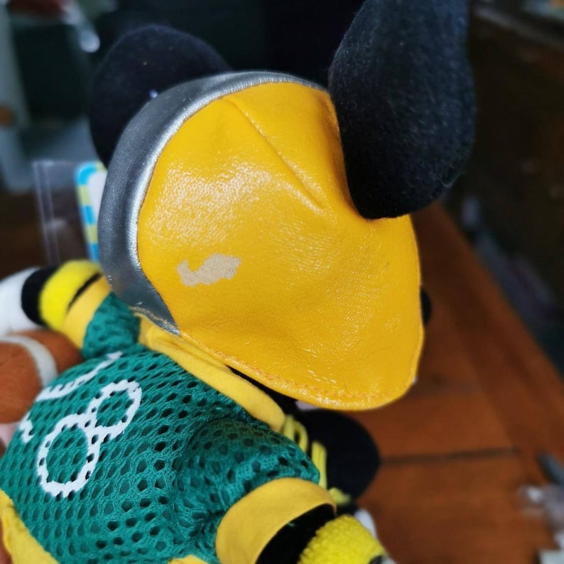 May include: A plush toy with a black and yellow head, wearing a green and yellow jersey with the number 8 on it. The toy has a silver helmet with a yellow visor.