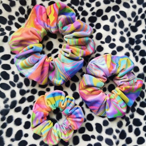 Op de afbeelding: Drie kleurrijke scrunchies met een levendig, wervelend patroon in roze, geel, blauw en groen. De scrunchies zijn gemaakt van een zachte stof en zijn geplaatst op een zwart-witte gespikkelde achtergrond. Haaraccessoires.