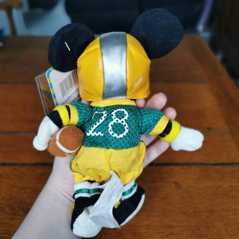 May include: A plush Mickey Mouse toy wearing a yellow and green football uniform with the number 28 on the front. He is holding a brown football and has a silver helmet on his head.
