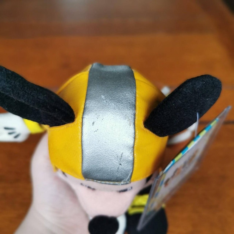 May include: A yellow and silver plush toy with black ears and a white face. The toy is wearing a yellow and silver helmet. The toy is a character from the Disney movie, "Planes".