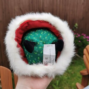 K&ouml;nnte beinhalten: Ein gr&uuml;nes Pl&uuml;schtier mit wei&szlig;em Pelzbesatz und roter Innenausstattung. Das Spielzeug hat ein Muster aus Stechpalmenbl&auml;ttern und schwarze Ohren. Das Spielzeug hat ein Etikett mit der Aufschrift "DN 20066".