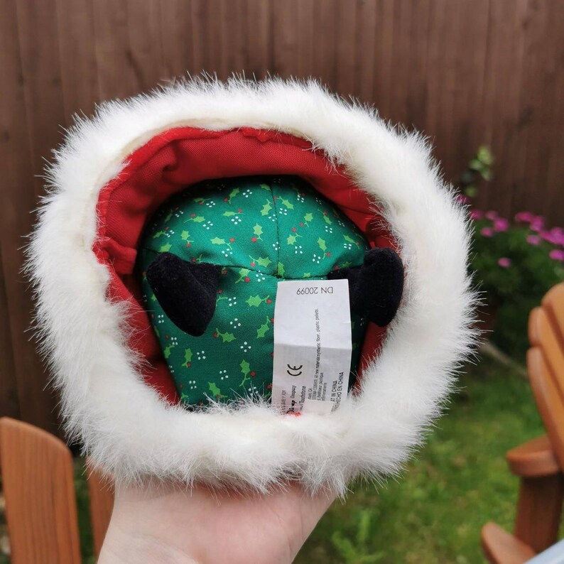 K&ouml;nnte beinhalten: Ein gr&uuml;nes Pl&uuml;schtier mit wei&szlig;em Pelzbesatz und roter Innenausstattung. Das Spielzeug hat ein Muster aus Stechpalmenbl&auml;ttern und schwarze Ohren. Das Spielzeug hat ein Etikett mit der Aufschrift "DN 20066".