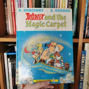 Könnte beinhalten: Ein Vintage-Asterix-Comic mit dem Titel "Asterix und der Zauberteppich" von René Goscinny und Albert Uderzo. Das Cover zeigt eine farbenfrohe Illustration von Asterix und seinen Freunden, die auf einem Zauberteppich fliegen.