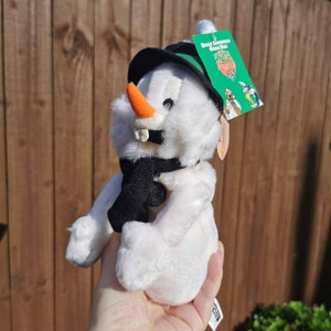 Könnte beinhalten: Weißer Plüsch-Schneemann mit schwarzem Schal, grünem Hut und orangefarbener Karottennase. Der Schneemann hält ein schwarzes Objekt in der Hand. Das Etikett lautet "Duck Dodgers Bean Bag".