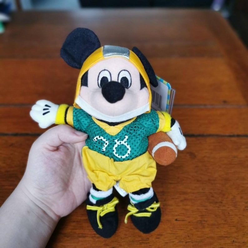 May include: A plush Mickey Mouse wearing a green and yellow football uniform with the number 78 on the jersey. He is holding a brown football in his right hand.