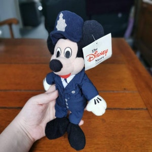 K&ouml;nnte beinhalten: Ein blaues Pl&uuml;sch-Mickey-Mouse-Spielzeug, das einen blauen Polizeihut und eine Uniform tr&auml;gt. Das Spielzeug hat ein Etikett mit der Aufschrift "The Disney Store".