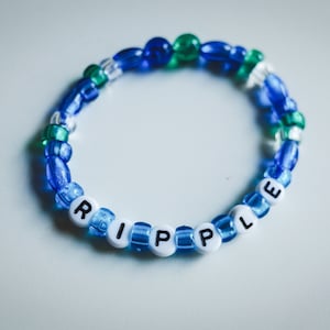 May include: A beaded bracelet with the word "RIPPLE" spelled out in white letter beads. The bracelet features blue, green, and clear beads. The bracelet is a handmade accessory.