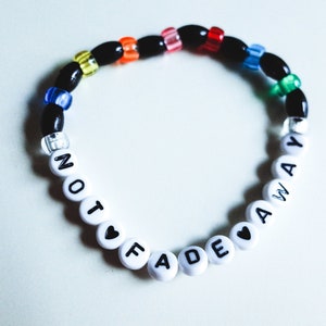 May include: A white beaded bracelet with the words "NOT FADE AWAY" spelled out in black lettering. The beads are interspersed with colorful beads in shades of red, orange, yellow, green, blue, and purple.
