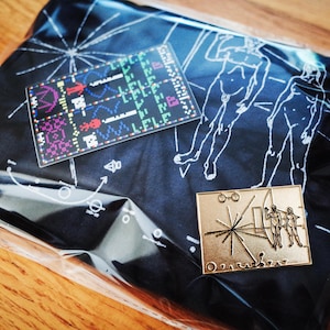 May include: A black fabric pouch with a white design of a starburst and figures. The pouch is packaged in a clear plastic bag. The pouch also features a gold metal pin with a starburst design and the text "Golden Record."
