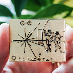 May include: Gold-colored metal pin featuring a stylized image of the Voyager Golden Record, a record containing sounds and images of Earth, sent into space on the Voyager spacecraft. The record is surrounded by a series of lines and circles, and includes a depiction of a man and woman.