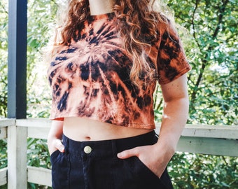 May include: A cropped tie-dye t-shirt in shades of orange and black with a spiral pattern. The shirt is worn by a person with long curly hair.