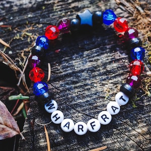 May include: A beaded bracelet with red, blue, and black beads, along with a star-shaped bead. The bracelet spells out the word "WHARFRAT" with white letter beads. The bracelet is resting on a weathered wooden surface.