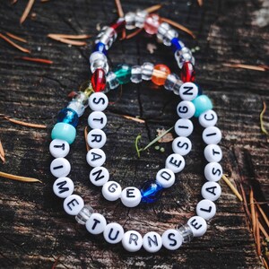 May include: Two beaded bracelets with the words "STRANGE THINGS DESIGN" and "ELASTIC TIME TURNS" on a wooden surface.