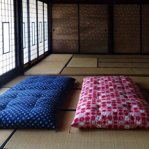 May include: Two rectangular floor mattresses placed side by side on a straw mat floor. One mattress is dark blue with a white circular pattern, and the other is pink with a white and red floral pattern. Both feature tufted stitching details.