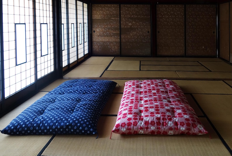 May include: Two rectangular floor mattresses placed side by side on a straw mat floor. One mattress is dark blue with a white circular pattern, and the other is pink with a white and red floral pattern. Both feature tufted stitching details.