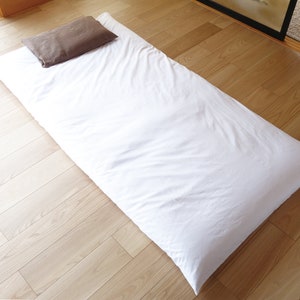 以下が含まれることがあります： 明るい木製の床に、白い布団マットレスと茶色の枕があります。マットレスは長方形で、柔らかく通気性のある生地で作られているようです。枕は長方形で、無地の茶色です。