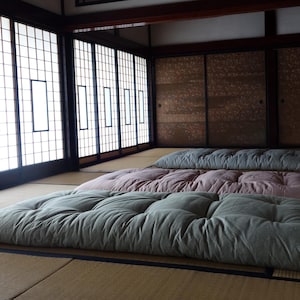 Puede incluir: Tres futones de suelo en una habitación tradicional japonesa. Los futones son en tonos verdes y rosas, dispuestos sobre un suelo de esterilla tatami. La habitación tiene puertas de papel y puertas correderas.