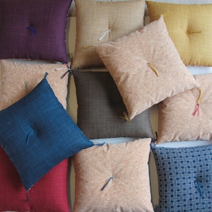 Puede incluir: Una colección de nueve cojines cuadrados en varios colores y patrones, incluyendo beige, marrón, azul, rojo y amarillo. Los cojines están hechos de una tela suave y texturizada y tienen un diseño japonés tradicional.