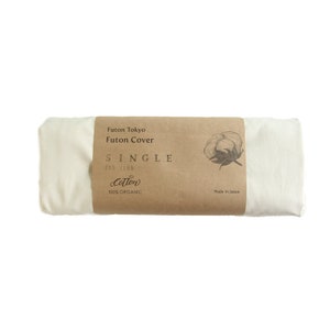 Op de afbeelding: Een opgerolde, crèmekleurige katoenen futonhoes met een bruine papieren band. De band bevat de tekst "Futon Tokyo Futon Cover SINGLE 205 X 105 Cotton 100% ORGANIC" en een katoenplantillustratie. Gemaakt in Japan.