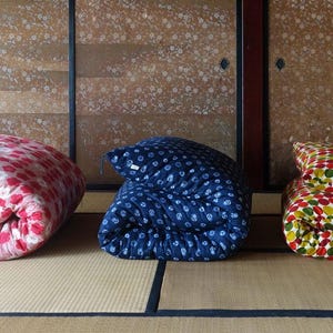 May include: Three folded futons arranged in a row. The left futon is pink with a white floral pattern. The middle futon is dark blue with a white geometric pattern. The right futon features a multi-colored circular pattern in red, green, and yellow.
