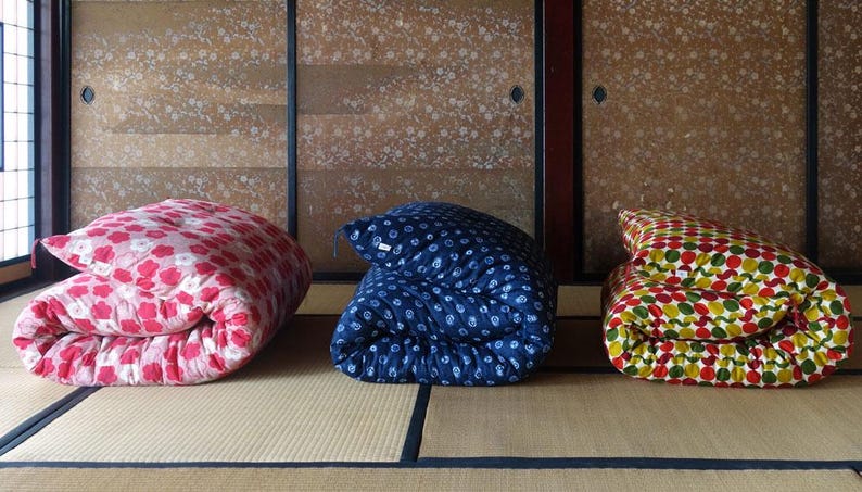 May include: Three folded futons arranged in a row. The left futon is pink with a white floral pattern. The middle futon is dark blue with a white geometric pattern. The right futon features a multi-colored circular pattern in red, green, and yellow.