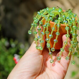 May include: A miniature terracotta pot holds a small plant with cascading strands of green beads. The beads are a mix of light and dark green, with some iridescent accents. The plant is held in a hand.