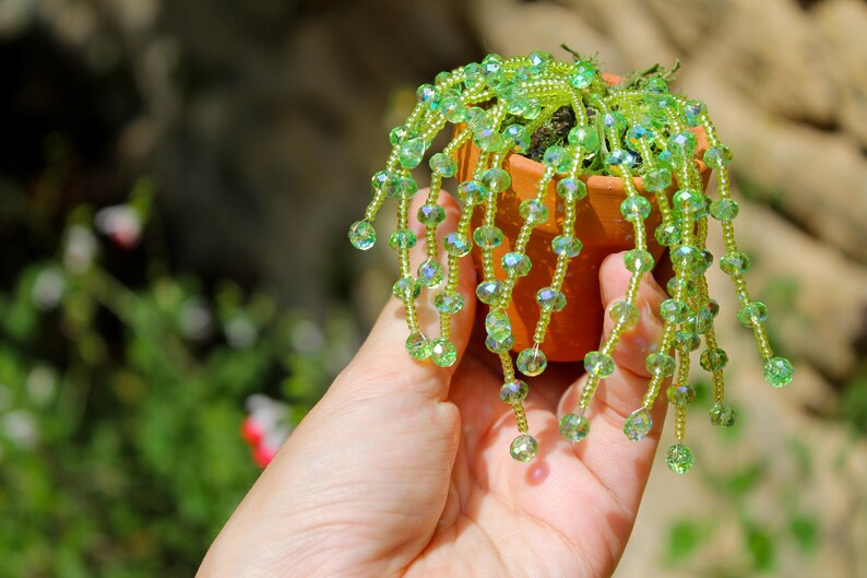 May include: A miniature terracotta pot holds a small plant with cascading strands of green beads. The beads are a mix of light and dark green, with some iridescent accents. The plant is held in a hand.