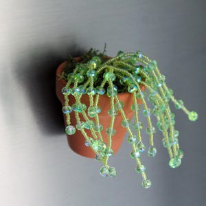 May include: A small terracotta pot with a beaded plant. The plant features strands of green and iridescent beads, resembling a cascading vine. The pot is attached to a gray surface.