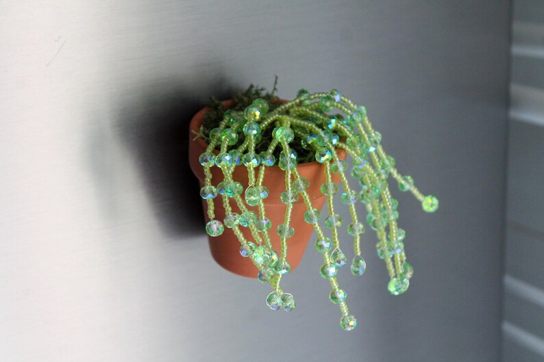 May include: A small terracotta pot with a beaded plant. The plant features strands of green and iridescent beads, resembling a cascading vine. The pot is attached to a gray surface.