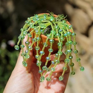 May include: A miniature terracotta pot holds a handcrafted beaded plant. The plant features strands of green beads, resembling cascading foliage. The beads have a faceted design, catching the light. The pot is held in a hand, with a blurred natural background.