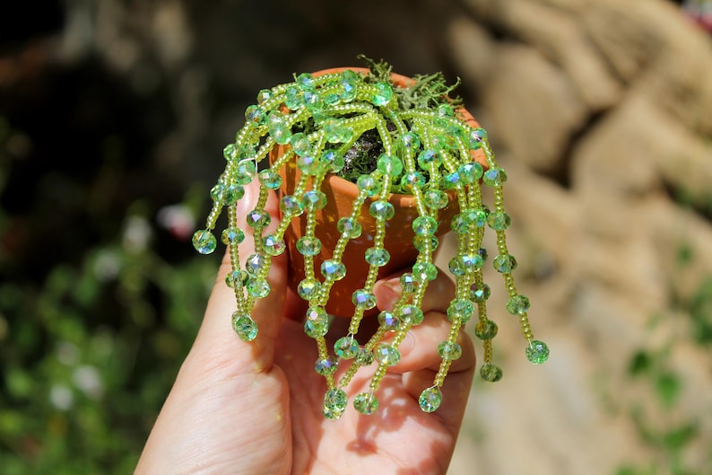 May include: A miniature terracotta pot holds a handcrafted beaded plant. The plant features strands of green beads, resembling cascading foliage. The beads have a faceted design, catching the light. The pot is held in a hand, with a blurred natural background.