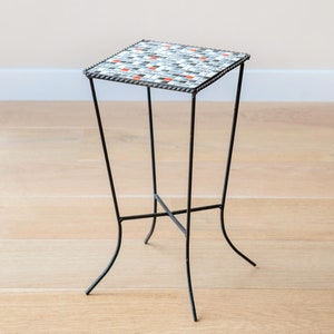 Op de afbeelding: Een kleine zwarte metalen bijzettafel met een vierkant mozaïekblad. De tegels zijn wit, zwart en oranje.