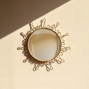Puede incluir: Un espejo redondo dorado con un marco ornamental en forma de sol. El marco presenta dise&ntilde;os delicados y arremolinados alrededor del espejo circular. El espejo est&aacute; montado en una pared de color claro, proyectando sombras.