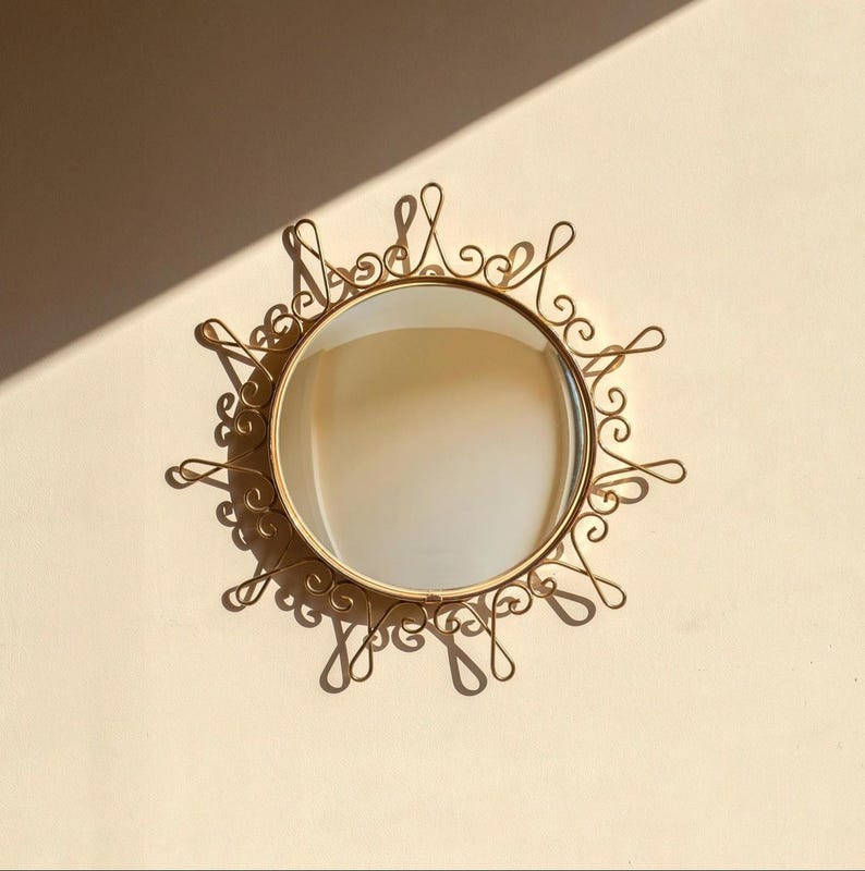 Puede incluir: Un espejo redondo dorado con un marco ornamental en forma de sol. El marco presenta dise&ntilde;os delicados y arremolinados alrededor del espejo circular. El espejo est&aacute; montado en una pared de color claro, proyectando sombras.
