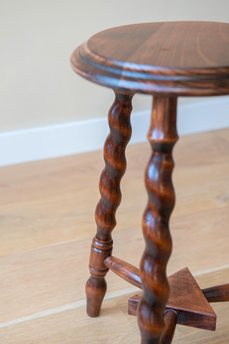 May include: A round wooden stool with a dark brown finish, featuring three distinctively twisted, spiral-shaped legs that connect to a central triangular support base. The stool has a smooth, circular top with a slightly rounded edge profile.