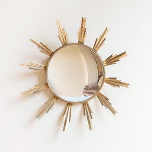 May include: A gold-coloured sunburst mirror with a round, reflective centre. The mirror's frame has a textured, beaded edge and radiating, stylised sun rays in varying shades of gold and silver. This decorative wall mirror adds a touch of vintage charm.