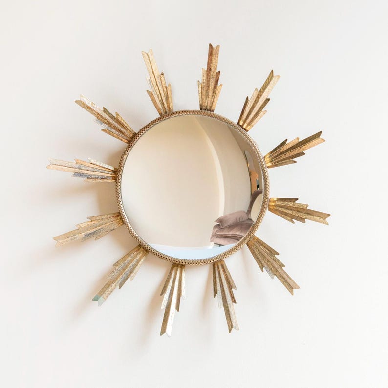 May include: A gold-coloured sunburst mirror with a round, reflective centre. The mirror's frame has a textured, beaded edge and radiating, stylised sun rays in varying shades of gold and silver. This decorative wall mirror adds a touch of vintage charm.