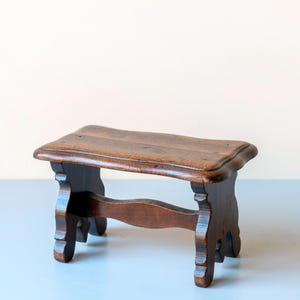 Op de afbeelding: Een kleine, antieke houten kruk met een donkerbruine afwerking. De kruk heeft een rechthoekige zitting met een gebogen rand en vier sierlijke poten. De poten zijn verbonden door een gebogen ligger. Afmetingen: ongeveer 30 cm lang, 20 cm breed en 25 cm hoog.