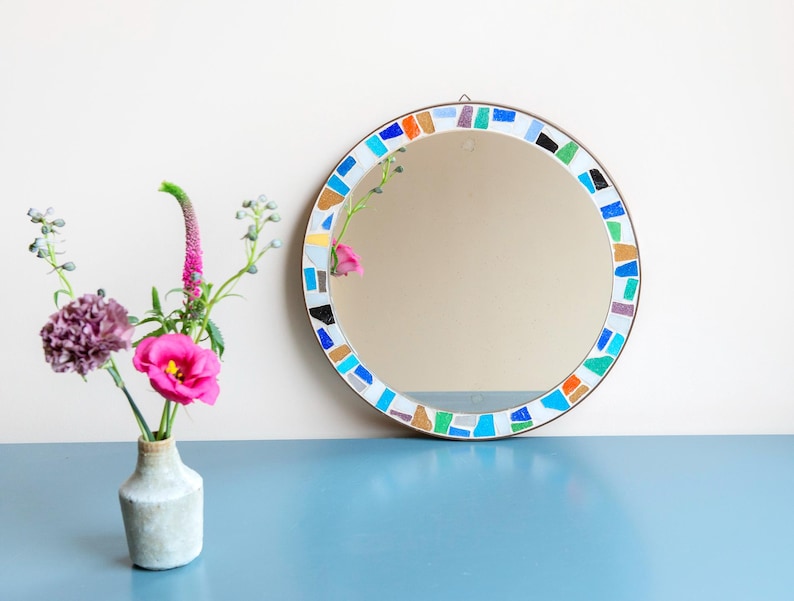 May include: A round mirror with a colorful mosaic border. The border is made of small, irregular pieces of glass in shades of blue, green, orange, and brown. The mirror is hanging on a white wall.