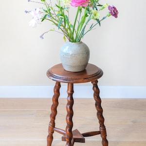 May include: A round wooden stool with three spiraled legs and a triangular support base, topped with a light gray ceramic vase containing a colorful arrangement of pink, purple, white, and green flowers with long, slender green stems.