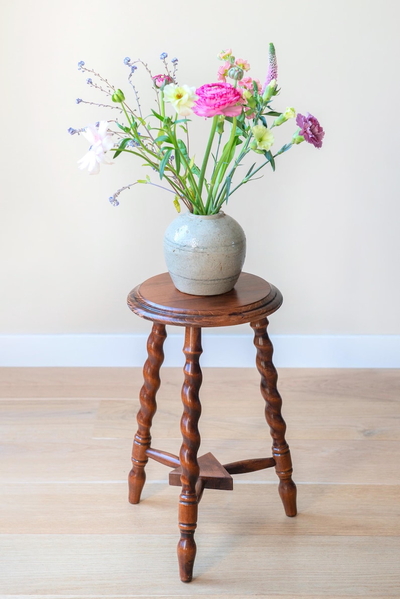 May include: A round wooden stool with three spiraled legs and a triangular support base, topped with a light gray ceramic vase containing a colorful arrangement of pink, purple, white, and green flowers with long, slender green stems.