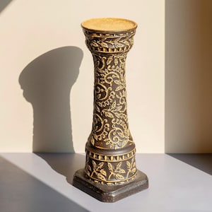 Op de afbeelding: Een donkerbruine decoratieve sokkel met ingewikkelde goudkleurige bloemen- en krulpatronen. De sokkel heeft een vierkante basis, een cilindrisch lichaam en een ronde bovenkant. Het ontwerp doet denken aan klassieke architectuur.