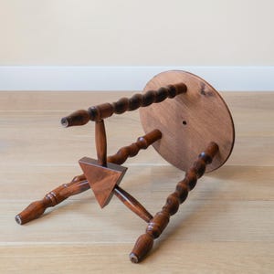 May include: A small wooden stool with a circular top and three legs featuring a twisted, spiral-turned design. The legs are connected by a central triangular wooden brace. The stool is finished in a medium-brown wood stain with visible grain.