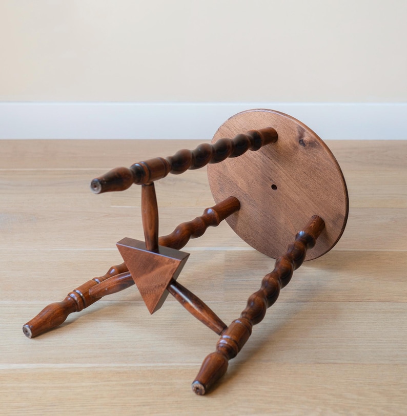 May include: A small wooden stool with a circular top and three legs featuring a twisted, spiral-turned design. The legs are connected by a central triangular wooden brace. The stool is finished in a medium-brown wood stain with visible grain.