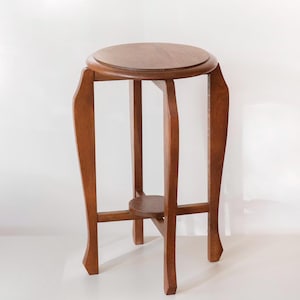 May include: A medium-brown wooden plant stand featuring a circular top surface, three curved legs that extend outward, and a smaller circular support shelf positioned centrally between the legs near the base of the structure.